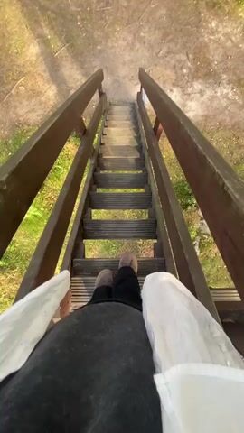 This is a first-person perspective video of someone descending a wooden outdoor staircase. The viewer's feet are visible at the bottom of the frame, wearing dark shoes and dark pants. They are wearing a white garment draped over their legs and a black garment on their upper body. The staircase has dark wooden railings on both sides. The steps are made of dark material with visible gaps. The surrounding environment appears to be a natural setting with dirt, dry leaves, and some green grass and foliage visible on either side of the stairs. The sunlight suggests it is daytime. The person is moving downwards, indicated by the changing perspective of the stairs and the ground below.
