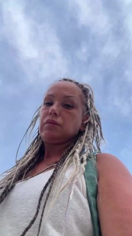The video shows a low-angle view of a woman with light brown skin and blonde braids, wearing a white tank top and a light green backpack. She looks directly at the camera with a serious expression. The sky is visible behind her, with a mix of blue and white clouds. The wind is blowing, causing her braids to move around her face. As the camera angle shifts slightly, a glimpse of buildings and trees appears in the background.
