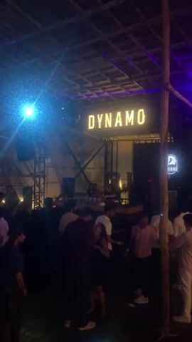 This media file depicts a lively scene at a venue called "DYNAMO" in Vagator, India. The setting appears to be an outdoor or semi-outdoor establishment, possibly a club or bar, with a rustic, possibly bamboo-constructed roof.

The main subjects are a group of people gathered in what seems to be a social or entertainment event. Several individuals are visible in the foreground and mid-ground, some engaged in conversation, others appearing to be dancing or observing. The lighting is dim and atmospheric, with bright spotlights, including a prominent blue light on the left, illuminating parts of the scene. The overall mood seems to be energetic and festive, suggesting a night out.

The most prominent visible text is the large, illuminated sign spelling out "DYNAMO" above the central area. There is also a circular logo with the name "DYNAMO" on a sign to the right. Other text elements, such as what might be branding on glasses or bottles, are visible on a bar counter in the mid-ground but are not clearly legible.

The time of day is clearly night, given the artificial lighting and the atmosphere. There are no strong indicators of weather, but the roof suggests protection from elements. The presence of a DJ booth or setup in the mid-ground, with individuals behind it, indicates that music and entertainment are being provided. The clothing of the people suggests a casual, modern attire suitable for a night club or beachside venue, common in locations like Vagator. The expressions and body language of the people suggest a relaxed and enjoyable atmosphere.