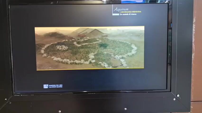 The video displays a digital screen with an image of a circular ancient settlement, surrounded by a stone wall and featuring several huts within. The background shows a misty landscape with hills and more structures. The screen also has text overlays. At the top right, the word "AQUINUM" is visible, followed by "L'ARCHAEOLOGIA PREVENTIVA" and "Un metodo di ricerca". At the bottom left, a logo with "MINISTERO PER I BENI E LE ATTIVITA CULTURALI" and "SOPRINTENDENZA ARCHEOLOGICA" is present. The video transitions to a new screen displaying text about the archaeological process, stating that individual testimonies (tombs, burials, traces of centuriation) are inserted into a single scheme to reconstruct lost scenarios. The text is presented in white against a dark background with a yellow border. The overall scene appears to be an exhibit or presentation, possibly in a museum or cultural institution. There are no people visible in the video.