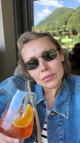 A woman wearing sunglasses and a denim jacket holds a glass of an orange beverage with ice and an orange slice, taking a sip through a straw. The background shows a window with a view of a mountain landscape.