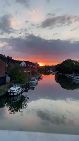 This video shows a sunset over a river with boats docked on either side. The sky is filled with clouds, some of which are illuminated by the setting sun with shades of orange, pink, and purple. The sun itself is low on the horizon, peeking through the clouds. The water reflects the colors of the sky and the surrounding buildings and trees. There are at least three boats visible in the foreground, two docked on the left side of the river and one on the right. The buildings appear to be houses or apartments, with some trees and greenery interspersed. The overall scene is peaceful and serene.