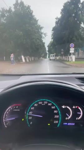 The video, shot from the perspective of a driver inside a car, shows a view through the windshield of a wet, tree-lined road on an overcast day. The car's dashboard is prominently displayed in the foreground, showing the speedometer reading around 20 km/h and the odometer at 89558. Outside, a white van is visible driving ahead on the road. The road appears to be a street in a town or village, with buildings and some pedestrians visible on the left side. Raindrops are visible on the windshield.