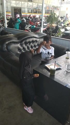 This media file shows two young children, likely siblings, in an outdoor seating area. The child on the left, a boy, is wearing a white t-shirt and is seated on a black tufted couch, looking at the child on the right. The child on the right, a girl, is wearing a black hijab and dress, and pink sandals. She is holding sunglasses in one hand and a piece of food in the other. She is looking towards the camera with a slight smile.

The setting appears to be a cafe or restaurant with an outdoor seating area. Behind the children, there is a black table with a drink in a plastic cup and a tablet or book. In the background, there is a large display of motorcycles, suggesting the location might be a business that sells motorcycles or a cafe frequented by motorcycle riders. The overall lighting suggests it's daytime, likely not too early or late. The ground is a concrete surface. There is a visible AC unit mounted on a building in the background. The scene is set in Matangjareung, Indonesia. The children appear happy and relaxed, enjoying their time together.
