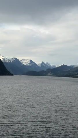 The video shows a wide body of water, likely a fjord or a lake, with snow-capped mountains in the background under a cloudy sky. The water has a rippled surface, suggesting a slight breeze. As the camera moves, the structure of a cruise ship becomes visible on the right side of the frame. The overall tone is serene and emphasizes the natural landscape.