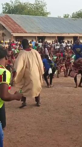 The video shows a group of people gathered in an open area, likely a courtyard, in front of a building with a corrugated iron roof. In the foreground, a few adults stand watching children perform a dance. The children are holding small flags and are dressed in what appears to be traditional clothing. Some of the adults are also dressed in traditional attire, including a man in a large beige robe who is dancing along. The crowd is watching the performance. The ground is dirt, and there are trees visible in the background.