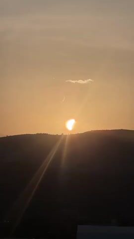 The video shows a time lapse of a sunset over a hilly landscape. The sun is setting behind the hills, casting a warm orange glow across the sky. There is a thin crescent moon visible in the sky above and to the left of the sun. A small, wispy cloud is also present near the moon. The foreground is dark and silhouetted against the bright sky, with what appears to be a building or structure visible at the bottom of the frame. The sun's rays are visible as bright streaks of light extending from the sun.