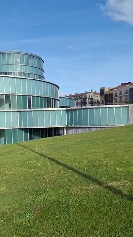 The video shows a modern building with a circular design. The building appears to be constructed primarily of glass and white metal, with a multi-tiered structure topped by a cylindrical tower. In front of the building is a grassy hill, and a shadow of the person filming the video is visible on the grass. In the background, there are other buildings with more traditional architectural styles. The sky is blue with some scattered clouds. The light is bright and the day appears sunny.