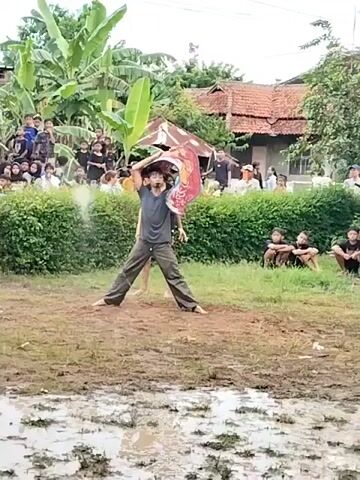The video shows a muddy field in a rural setting. A person wearing a gray shirt and dark pants is performing some kind of dance or theatrical performance. They have a large, colorful object balanced on their head. There is a small audience of people watching, many of whom appear to be young. In the background, there are houses and trees. The ground is wet and muddy, with standing water visible in places. Some children are sitting on the grass to the right of the muddy field.