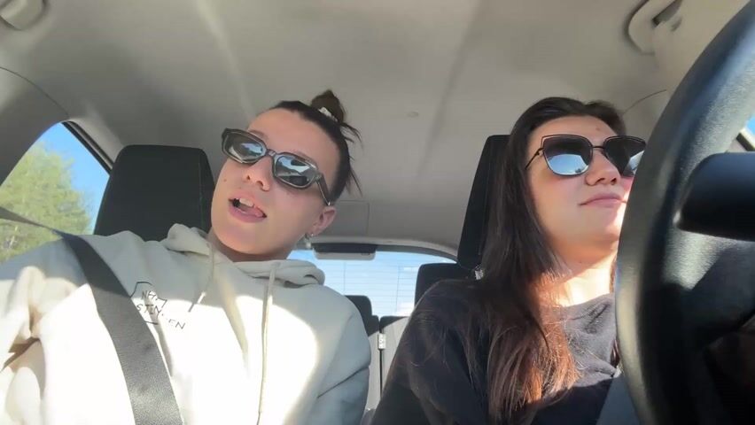 Two women are in a car. One is driving. They are both wearing sunglasses. The driver has long dark hair, and is wearing a black shirt. The passenger has her hair in a bun. She is wearing a light colored hoodie. Both women are looking forward. The sky is visible through the windshield. The passenger leans her head to the side. The driver makes a hand gesture while talking. Music is playing.