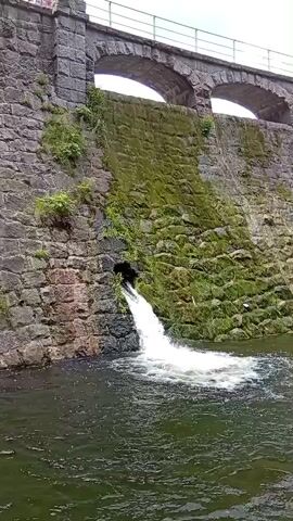 A stone dam with arched structures on top is visible. A pipe protrudes from the side of the dam, releasing a stream of water that creates foamy turbulence in the dark water below. The dam's stone surface is partially covered in green moss. The sky above is overcast.
