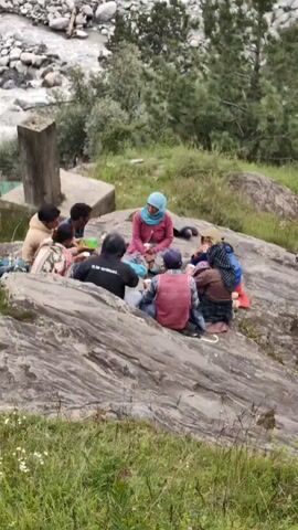 A group of seven people are sitting in a circle on a large rock formation. The group consists of at least two adults and five children, with some individuals wearing head coverings. They appear to be having a meal or snack, with cups and food items visible. The surrounding environment is a mountainous landscape with a river or stream in the background, trees, and grassy slopes. The weather appears to be overcast. The video pans out to show a wider view of the valley and mountains.