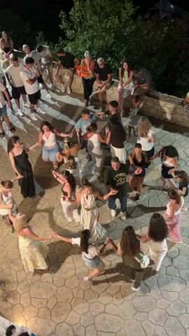 A high-angle shot shows a group of people holding hands in a circle and dancing. They are on an outdoor paved area with hexagonal stone tiles. The background is dark, suggesting nighttime, with some foliage visible at the top of the frame. Some people are standing around the perimeter watching the dancers. The people dancing are wearing casual summer clothing, including dresses, shorts, and t-shirts. The atmosphere appears festive.