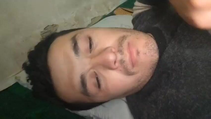 A man lies on his back, on a bed with a pillow and a green and white patterned blanket. He looks directly at the camera and rests his hand on his face. The man is wearing a dark gray sweater. The background is out of focus and shows a white wall.