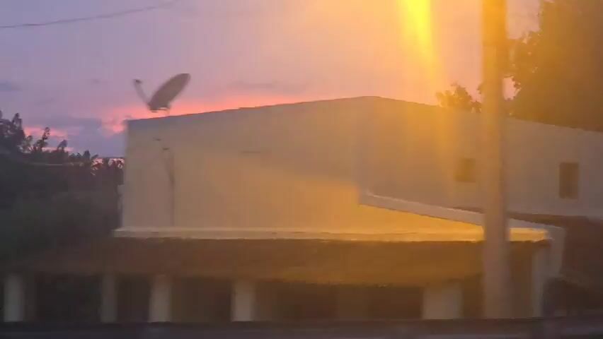 A video captures an outdoor scene during twilight. The first few seconds show the side of a building with small, dark windows. The camera then pans to a multi-story building with an external staircase and a balcony. The sky has hues of pink and purple, indicating sunset or sunrise. In the subsequent frames, the video shows trees and foliage blurring as if in motion. The final seconds reveal a wider landscape with a vibrant, colorful sky streaked with orange and pink. In the distance, there are buildings and some vegetation. There are no people visible in the video.