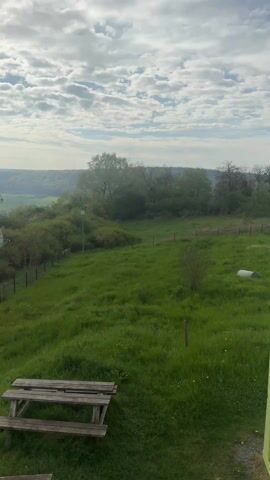 The camera is positioned in an elevated location overlooking a grassy field.  In the foreground is a wooden picnic table.  The field, which contains a scattered number of saplings, is bordered by a black wire fence.  On the other side of the fence are bushes and trees.  A pole, with what appears to be two satellite dishes and an antenna attached, is visible in the middle distance.  Beyond the trees and bushes is a valley vista of hazy green hills.  The sky is cloudy, with patches of blue visible.