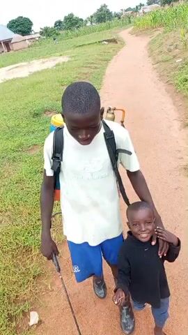A video shows two young boys standing on a dirt path. The taller boy, on the left, is wearing a white t-shirt, blue shorts, and black rubber boots. He has a black backpack sprayer on his back with a yellow tank and a metal wand in his right hand. The shorter boy, on the right, is wearing a black shirt, blue shorts, and brown sandals. He has his left arm around the taller boy's waist. Both boys are looking at the camera and smiling. The path is lined with dry grass and some green vegetation. In the background, there are trees and some small buildings. The sky is overcast.