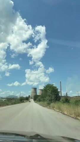 The video begins with a view from inside a vehicle, looking forward through the windshield. A paved road stretches ahead, leading towards a distant industrial complex. The sky is a vibrant blue, dotted with fluffy white clouds. On either side of the road, there is dry grass and vegetation, with some trees visible. In the distance, two large, distinctive cooling towers of a power plant are prominent, with plumes of steam or smoke rising from their tops. A tall smokestack is also visible. A few cars are seen on the road. The perspective shifts slightly as the vehicle moves, offering a clearer view of the cooling towers and the surrounding landscape under a sunny sky.