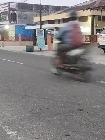 A street scene at dusk with buildings lining one side of the road. A person on a motorcycle is in motion in the foreground, blurred due to movement. Further down the road, a parked car and several other motorcycles are visible. People are seen walking and interacting near the buildings. The road surface appears to be asphalt with some cracks and wear.