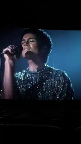 The video shows a movie screen in a dark cinema. The first scene on the screen features a young man singing into a microphone. He wears a sparkling shirt. The second scene shows a man in a white shirt and hat sitting at a table. In the third scene, a young man sits at a desk and writes in a notebook. The camera pans slightly around the room. The scene ends.