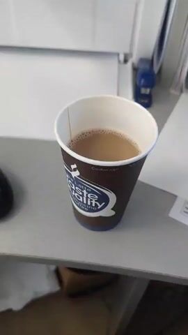 A close-up shot shows a paper coffee cup sitting on a gray surface. The cup is brown and has a white interior. The cup has a logo that reads "Taste Quality" with "Latte Cappuccino Coffee" underneath. There is a caution label "Caution: Hot!" on the side of the cup. Inside the cup is a light brown liquid, likely coffee with cream or milk. The background shows a white printer or similar office equipment and other office supplies such as a stapler and documents. The setting appears to be an office environment.