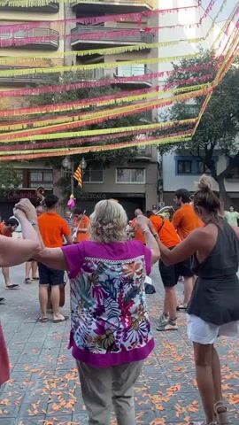 A group of people are dancing in a circle outdoors, holding hands. They are in a street decorated with colorful bunting strung between buildings. A band is playing music in the background, with several members wearing orange shirts. Some of the dancers are also wearing orange. There are orange confetti or streamers scattered on the ground. The buildings in the background appear to be apartment buildings. The overall atmosphere is festive.