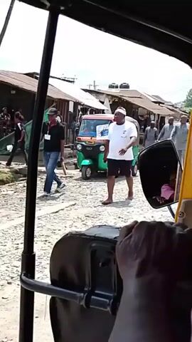 The video captures a street scene in Obunta-Isiala, Nigeria, from the perspective of someone riding in a yellow auto-rickshaw (Keke Napep). The vehicle appears to be moving slowly or is stationary.

Several individuals are visible on the unpaved, rough street. In the foreground, a man in a white cap, white t-shirt, and dark shorts walks towards the camera. Further down the street, another green and orange auto-rickshaw is visible. Several men in light-colored traditional Nigerian attire are walking away from the camera. One man in a red t-shirt is also seen in the background.

The setting consists of simple buildings, possibly shops or residences, with corrugated iron roofs lining the street. The time of day is clearly daytime, with bright sunlight indicating sunny weather. No specific emotions are discernible on the faces of the people, who appear to be going about their daily routines. No legible text is visible on any signs or clothing.