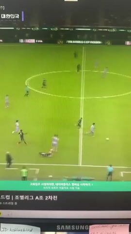 A computer monitor displays a live broadcast of a soccer match. The game is being played on a vibrant green field with white markings. Several players in purple and white uniforms are visible on the field. In the lower portion of the screen, a banner with Korean text indicates match information, including "JOINING LEAGUE A Round 2". The monitor itself is visible at the bottom, with a Samsung logo and some handwritten notes.