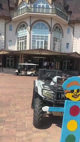 The video captures an outdoor scene in front of a large, ornate building, possibly a hotel or resort, with a grand entrance featuring multiple arched windows and a covered walkway. The time of day is daytime, with bright sunlight casting distinct shadows, suggesting fair weather.

In the foreground, a camouflage-patterned ATV is parked on a paved area constructed of interlocking bricks. Attached to the front of the ATV is a colorful, whimsical decoration resembling a traffic light with a cartoon face wearing a cap.

To the left of the ATV, a white golf cart is visible, suggesting recreational activities or transportation within the venue. Several people are gathered near the entrance of the building, some appearing to be guests or staff. Their casual attire and the relaxed setting imply a leisurely atmosphere.

A sign on the back of the ATV displays text in what appears to be Cyrillic script. The prominent text reads "ПРОКАТ КВАДРО..." which translates to "Quad Rental..." followed by what seems to be a phone number, "(999)". The overall scene suggests a location offering outdoor recreational rentals and accommodations. The cheerful decoration on the ATV adds a playful element to the otherwise formal architecture of the building.