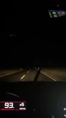 The video shows a car driving on a dark, empty road at night. The dashboard is visible, showing a speed of 95, 96, and 97, with the gear in D5. The headlights illuminate the road ahead, marked with white lines and roadside reflectors. A phone is mounted above the dashboard. The environment appears to be rural with flat, dark fields on either side of the road.