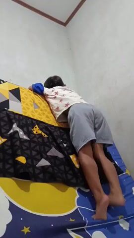 A young child with dark hair, wearing a white t-shirt with a red star pattern and light blue trim, and grey shorts, is lying on a bed with a patterned blue and yellow bedsheet. The child is positioned with their back to the camera, leaning towards the headboard of the bed. The bedsheet features yellow stars and crescent moon shapes. The child's legs are extended, and their feet are visible. The child's hands are resting on the headboard, which has a yellow and black geometric pattern. The setting appears to be indoors, with a plain, light-colored wall visible in the background. The lighting suggests it could be daytime, but it is difficult to determine the exact time. The child's posture and relaxed demeanor do not convey any strong emotions. There is no visible text in the media file.
