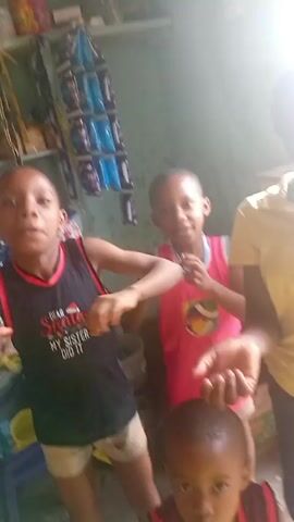 Four children stand indoors. The child in the center wears a pink shirt and white shorts. To his left is a child in a dark blue tank top with white shorts. The tank top has a graphic that reads "DEAR SANTA MY SISTER DID IT." The child at the front is wearing a maroon tank top. To the right stands an adult wearing a yellow shirt. The children are moving their arms. There are packages hanging on the wall behind the children.