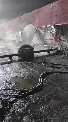 A water sprinkler is attached to a mechanical device with a large disc, possibly a saw, that is spraying water into the air. The area is wet and muddy with puddles of water. In the background, there is a truck and two people wearing yellow vests, one of whom appears to be operating the sprinkler. There is a large tarp or netting structure behind the equipment. The scene is illuminated by bright lights, suggesting it is nighttime.