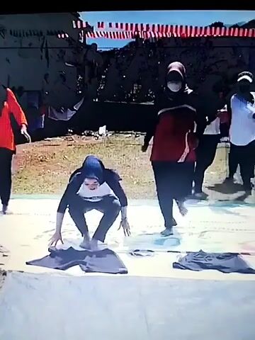 A group of people are outdoors on mats on the ground. The person in the center is wearing a blue headscarf and a grey and white long-sleeved shirt with writing on it. They are in a squatting position on the mat, with black pants spread out in front of them. To their right, a person in a red and black shirt is standing. To their left, a person in an orange shirt is also on a mat. The ground around the mats is covered in grass and dirt. In the background, there is a dark structure with red and white decorations strung across the top. The sky is blue and appears to be daytime.