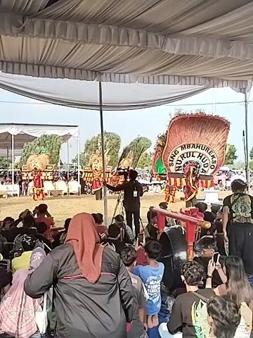 This is a video clip taken at an outdoor event in Plosorejo, Indonesia. The scene is set under a large white tent, providing shade for the spectators and performers. In the foreground, a crowd of people are seated on the ground, facing a performance area. Many in the crowd are holding up their phones, suggesting they are recording or taking pictures of the event.

The performance area in the middle ground features several individuals in colorful costumes, some with elaborate headdresses that resemble peacock feathers. There are also large, decorated floats or structures, one of which has text on it that reads "URSING MBAHUREKO NURUL HUDA". In front of these performers, a person dressed in black is standing with a camera on a tripod, seemingly filming the event.

Further in the background, more people are visible, and there are structures that appear to be stages or platforms. The overall atmosphere seems festive and engaging, with the audience actively participating by documenting the performance. The lighting suggests it is daytime, likely late morning or afternoon, with bright, natural light. The weather appears to be clear, as evidenced by the bright sky. The event is likely a cultural performance or festival, given the traditional costumes and elaborate decorations.