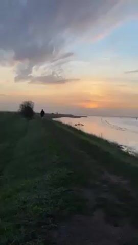The video depicts a man walking away from the viewer on a grassy path next to a body of water at sunset. The sky is filled with clouds tinged with shades of pink, orange, and yellow as the sun sets on the horizon. To the left of the path is a field of green grass with a small bush. The path itself is dirt and grass. The body of water appears calm, reflecting some of the colors of the sunset. The man is silhouetted and appears to be alone.