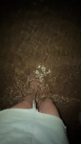 A person stands at the edge of the water at night. They are wearing denim shorts, and the water comes up to their shins. They shift their weight from foot to foot, causing ripples in the water. The sand on the beach is visible through the water.
