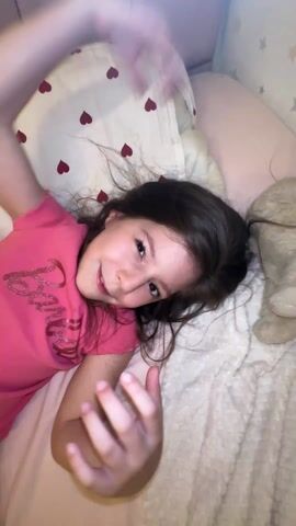A young girl lies on a bed with a pillow decorated with red hearts. She is wearing a pink shirt with a Barbie logo. An adult's hand waves in front of the camera and the girl. The camera is unsteady, and the shot is close. The girl seems playful. The room lighting is soft and warm.