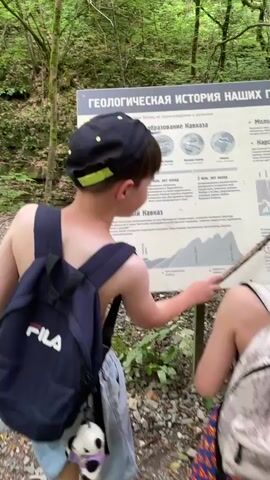 In a lush, green forest setting, likely Izmaylovka, Russia, a group of people are gathered around an informational sign. The sign, titled "GEOLOGICAL HISTORY OF OUR MOUNTAINS" in Russian, appears to be explaining the geological history and formation of the Caucasus mountains. Several individuals, including children and an adult woman, are looking at the sign. One child, wearing a black baseball cap with neon yellow accents and a Fila backpack, is pointing at the sign with a stick. Another child, wearing a pink baseball cap and a white bucket hat, is also looking at the sign, while the adult woman is holding a smartphone and seemingly reading something from it or taking a photo. The scene suggests an educational outing or nature walk. The time of day is unclear, but the presence of green foliage and dappled light suggests it's daytime, possibly overcast or shaded. The ground is covered with rocks and leaves, typical of a forest floor.