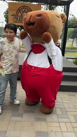 A boy poses in front of a building with two large teddy bear mascots. He is wearing a white T-shirt with a graphic print, blue jeans, and Adidas tennis shoes. The mascots are dressed in formal wear, one with a red dress and the other with red trousers and a white shirt. There is a wooden sign in the background that reads "PLAZA TRADICIÓN". A woman with a purple backpack walks past in the beginning of the video.