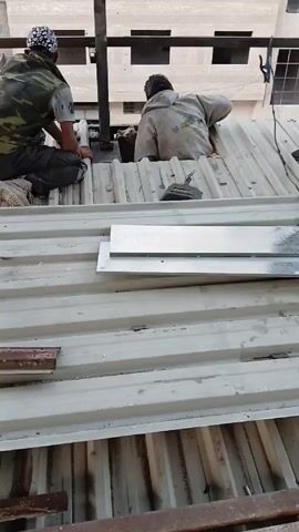 Two individuals are working on a corrugated metal roof. One is kneeling and appears to be working on something attached to a dark metal structure that protrudes from the roof. The other individual is leaning into an opening in the roof. Several rectangular pieces of metal are on the roof. The setting appears to be an urban environment.