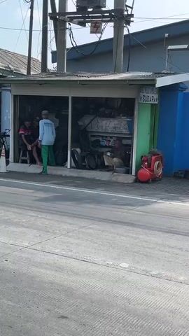 The video shows a street scene. In the foreground, the back of a motorcycle is visible, with a license plate reading "N 6488 BY". The background features a small shop, possibly a tire repair shop, with several people present. Inside the shop, there are tires and tools. A man in a light blue shirt and green pants stands in the doorway. Two other men are seated inside. Outside the shop, a rusty pickup truck drives by, with a man in the driver's seat. The truck has a spare tire in the bed. Overhead, electrical wires and poles are visible. The overall environment appears to be a roadside commercial area.