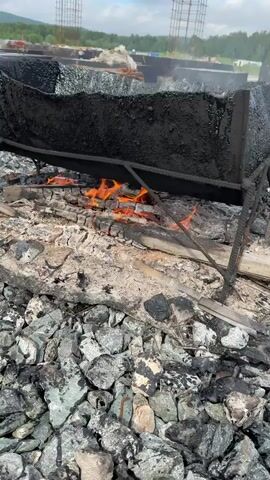 The video shows a black, tarp-like material that is contorted and melted, likely from intense heat. Below this material, a fire burns brightly, with orange flames licking at charred wooden logs and ash. The ground surrounding the fire is covered in grey and black gravel, interspersed with burnt debris. In the background, a construction site is visible. Several tall, rebar-reinforced concrete structures stand incomplete, suggesting the construction of a building or infrastructure. Piles of construction materials, including metal rods, are also present. The overall scene depicts an active fire at a construction site, possibly due to the melting or burning of construction materials.