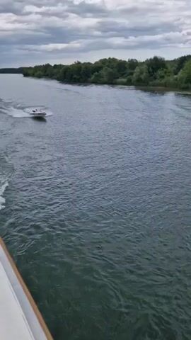 A white and blue speedboat with an outboard motor, carrying three individuals, travels left to right on a river. The boat creates a wake behind it. The river is surrounded by lush green trees and fields under a cloudy sky.