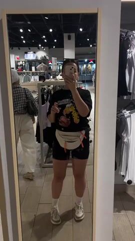 The media is a vertical, full-body mirror selfie of a young woman in a clothing store. She is standing in front of a large, framed mirror, taking the photo with her smartphone, which is held up to cover her face. The woman is wearing a black t-shirt with a graphic print featuring cartoon characters, black biker shorts, white socks, and light-colored sneakers. A white fanny pack with orange zipper pulls is worn across her waist. Her hair is pulled back in a bun, and she appears to be wearing sunglasses on top of her head.

In the background, the store is visible with clothing racks displaying various garments. To the left of the main subject, another person wearing a headscarf and a plaid shirt is browsing the racks. The store appears to be well-lit with overhead lighting, and the floor is tiled. The overall setting suggests a retail environment, possibly a department store or clothing boutique. The reflection in the mirror provides a clear view of the woman and her surroundings. There is no indication of the time of day or weather outside. The woman's pose and the selfie format suggest a casual and personal moment, perhaps while shopping or trying on clothes. There are no discernable emotions beyond a neutral or pleasant expression. No visible text is clear enough to be transcribed.