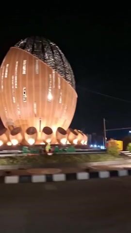 The video shows a large, egg-shaped structure at night. The structure is illuminated from within and by spotlights on the ground. It has a wooden-like exterior with vertical slits that allow light to escape. At the base of the structure, there are several smaller, rounded elements that are also lit from within. In front of the structure, large yellow letters spell out "SIGLI". The camera is moving, suggesting it is capturing footage from a vehicle. The surrounding area is dark, with only the illuminated structure and some streetlights visible.