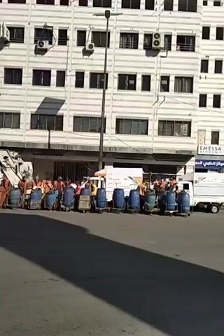 The video shows a group of sanitation workers, dressed in orange vests, standing behind a row of trash cans and blue barrels in a city street. The workers appear to be in a line, standing at attention. Behind them are several garbage trucks, one of which is yellow. The street is asphalt and has visible shadows. Tall buildings line both sides of the street. A silver car passes in front of the workers at one point. The sky is clear and blue.