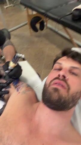 Here is a description of the video.

A man with a beard and a nose ring is lying down and getting a tattoo. He is shirtless. The tattoo is on his left shoulder. The tattoo artist is wearing black gloves and is using a tattoo gun. The room has tiled floors. There is a black massage table to the right.