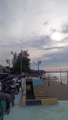 The video shows a waterfront scene with several motorcycles parked on a concrete walkway. A set of blue and white stairs leads down to a sandy beach with calm water. There are several wooden posts standing in the water. In the background, there are some buildings and trees, with a cloudy sky overhead. Two flags with a red and white pattern are visible, attached to poles. The sun is visible through the clouds, casting a soft light on the scene. The camera pans slowly from left to right, showing more of the beach and the water.