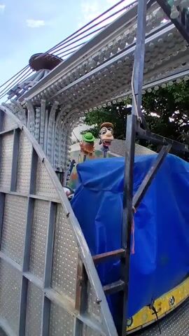 The video shows a carousel with a blue tarp covering the center. There are two figures visible on the carousel: a duck with a blue hat and a horse with an orange mane and tail. The carousel is surrounded by metal framework and lights. The background consists of trees and a cloudy sky. The video appears to be taken from a low angle, looking up at the carousel. The camera is moving slightly, as if someone is walking around the carousel.